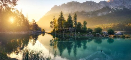 Foto de Lago idílico al amanecer, un pintoresco panorama con majestuosas montañas y la luz dorada del sol que entra detrás de los árboles - Imagen libre de derechos
