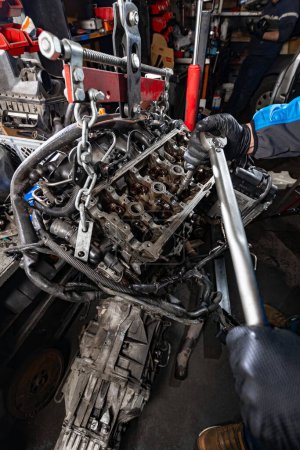 Experience auto expertise as the mechanic gloved hands skillfully manipulate a large wrench, loosening bolts on a suspended car engine, lifted from the chassis with a manual hoist for precise repair