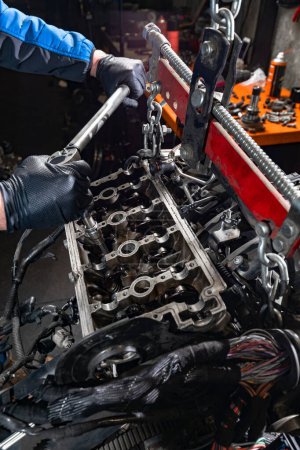Photo for Delve into the world of automotive mastery as the skilled hands of a mechanic, clad in protective gloves, meticulously handle a large wrench to loosen bolts on a car engine suspended from the chassis by a manual hoist, ensuring meticulous maintenance - Royalty Free Image