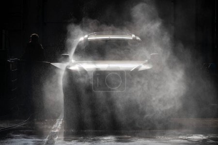 Man beobachte die Silhouette einer Person, die in einem Auto-Service-Center ein Auto mit einem Wasserstrahl wäscht. Die Szene wird durch Hintergrundbeleuchtung beleuchtet, die Wassertröpfchen werden hervorgehoben und ein dynamisches Ambiente geschaffen. Perfekt für automobile Konzepte.