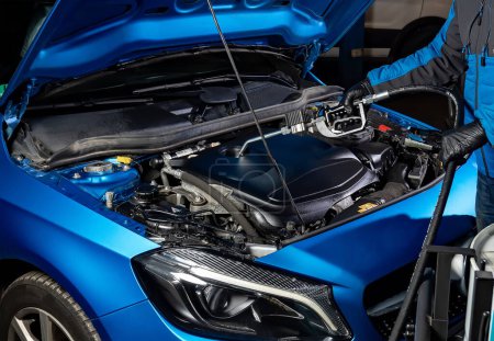 Kfz-Instandhaltung in Aktion als Mechaniker füllt den Motor eines blauen Autos durch eine geöffnete Motorhaube mit einer Ölpumpe, nachdem er das alte Öl entleert hat, Wechsel für optimale Motorleistung