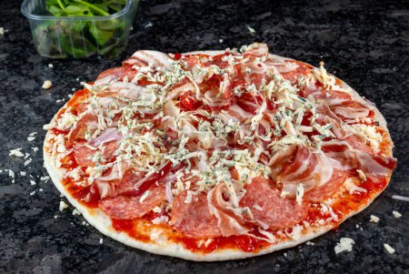 Téléchargez les photos : Voici une pizza alléchante ornée de jambon, de salami, de fromage, d'oignons et d'herbes fraîches, reposant sur un comptoir de cuisine en granit, attendant impatiemment son tour dans le four. L'anticipation se construit à mesure que les arômes salés remplissent l'air - en image libre de droit