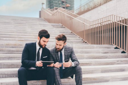 Photo for Two businessman sitting outdoors staircase using sharing tablet and smartphone discussing - Royalty Free Image