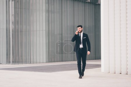 Photo for Young bearded elegant businessman walking outdoors talking smartphone - Royalty Free Image