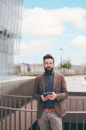 Téléchargez les photos : Jeune homme barbu à l'extérieur posant appareil photo tenant tablette ordinateur souriant serein et confiant - en image libre de droit