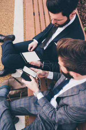 Foto de Dos elegantes hombres de negocios sentados en el banco al aire libre usando tableta y teléfono inteligente - Imagen libre de derechos