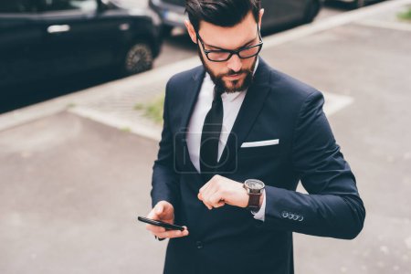 Photo for Young professional elegant bearded businessman holding smartphone checking time on wrist watch - Royalty Free Image