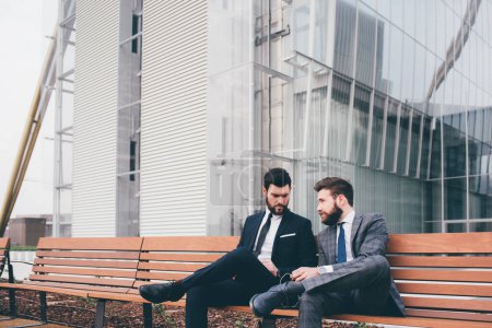 Foto de Dos jóvenes y elegantes hombres de negocios sentados al aire libre discutiendo y charlando - Imagen libre de derechos