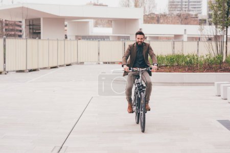 Foto de Joven hombre de negocios barbudo casual montar una bicicleta al aire libre en la ciudad- ecológico, transporte, concepto de negocio - Imagen libre de derechos