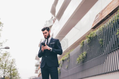 Téléchargez les photos : Jeune homme d'affaires élégant barbu à l'extérieur en utilisant la messagerie smartphone ou la lecture e-mail - en image libre de droit