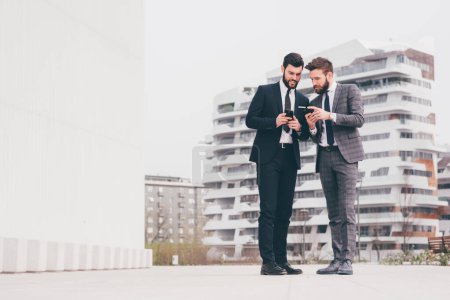 Téléchargez les photos : Deux jeunes hommes d'affaires contemporains en plein air dans la ville en utilisant le téléphone intelligent - entreprise, technologie, concept de communication - en image libre de droit