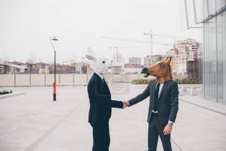 Téléchargez les photos : Poignée de main d'affaires devant un immeuble de bureaux entre deux hommes d'affaires contemporains portant des masques - carnaval ; Halloween ; concept étrange - en image libre de droit