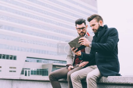Téléchargez les photos : Deux jeunes hommes d'affaires barbus assis en plein air en utilisant une tablette - en image libre de droit