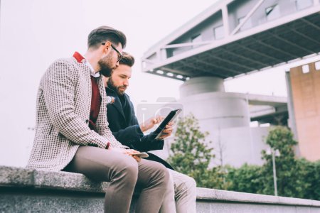 Photo for Two young bearded business men outdoor sitting using tablet - Royalty Free Image