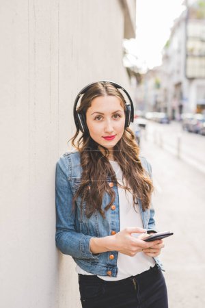 Foto de Joven hermosa mujer rubia caucásica escuchando música con auriculares y teléfono inteligente de la mano celebrar al aire libre en la ciudad, mirando en la cámara - música, tecnología, concepto sereno - Imagen libre de derechos