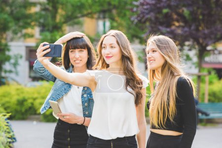 Foto de Tres jóvenes hermosas mujeres caucásicas jóvenes millennials, tomando selfie con la mano del teléfono inteligente celebrar al aire libre en la ciudad, sonriendo - red social, tecnología, concepto de amistad - Imagen libre de derechos
