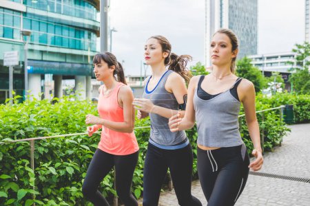 Téléchargez les photos : Trois jeunes belles femmes millénaires portant un survêtement courant en plein air dans la ville - jogging, coureur, concept de bonheur - en image libre de droit