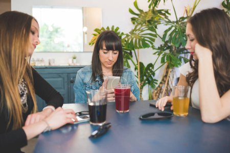 Téléchargez les photos : Trois jeunes femmes assis à l'intérieur bar bavarder en utilisant smartphone boire boisson gazeuse - en image libre de droit