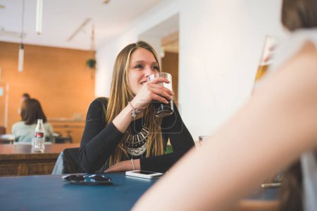 Téléchargez les photos : Deux jeunes femmes assises à l'intérieur bar bavarder et parler boire une boisson gazeuse - en image libre de droit