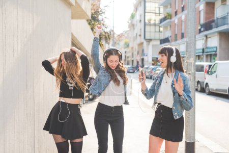 Foto de Tres hermosas jóvenes millennials escuchando música bailando divirtiéndose juntas - Imagen libre de derechos
