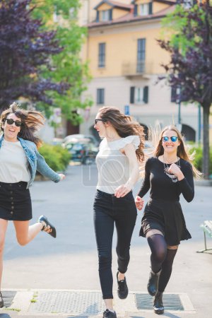 Foto de Tres mujeres jóvenes millennials al aire libre en la ciudad corriendo y saltando divirtiéndose sonriendo - Imagen libre de derechos