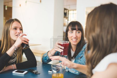 Téléchargez les photos : Trois jeunes femmes assises à l'intérieur bar bavarder et parler boire une boisson gazeuse - en image libre de droit