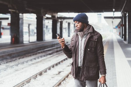 Téléchargez les photos : Jeune homme noir à l'aide d'un téléphone intelligent gare navette - en image libre de droit