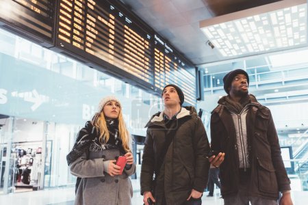 Foto de Tres amigos multiétnicos viajero de pie estación de tren comprobar el horario - Imagen libre de derechos