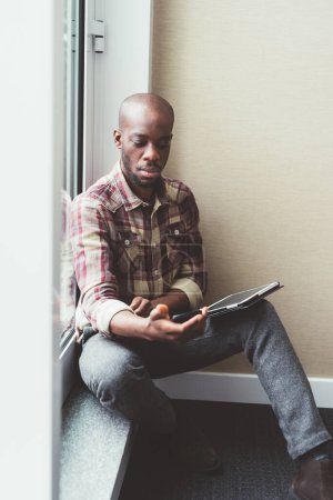 Photo for Black man sitting close to window and interacting with smartphone - Royalty Free Image