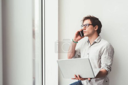 Téléchargez les photos : Jeune caucasien homme intérieur en utilisant ordinateur parler smartphone - en image libre de droit