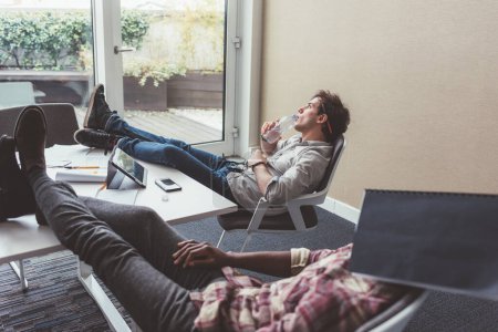 Foto de Dos hombres de negocios que se relajan en la oficina con piernas en la mesa durante el descanso - Imagen libre de derechos