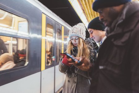 Téléchargez les photos : Trois jeunes amis adultes attendent métro et vérifier smartphone - en image libre de droit