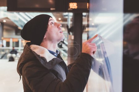 Téléchargez les photos : Jeune homme caucasien adulte debout à la gare et interagissant avec écran tactile - en image libre de droit