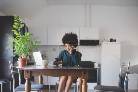 Téléchargez les photos : Jeune femme multiethnique à l'intérieur à la maison en utilisant l'ordinateur et smartphone télétravail - en image libre de droit