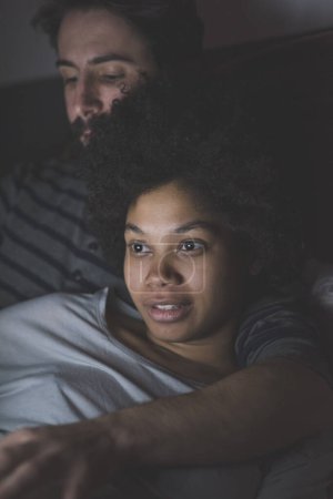 Foto de Joven pareja multiétnica abrazando dormitorio viendo tv - Imagen libre de derechos