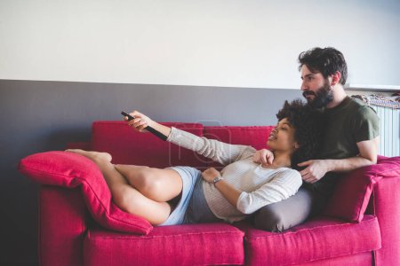 Téléchargez les photos : Jeune couple multiethnique à l'intérieur appartement assis canapé regarder la télévision - en image libre de droit