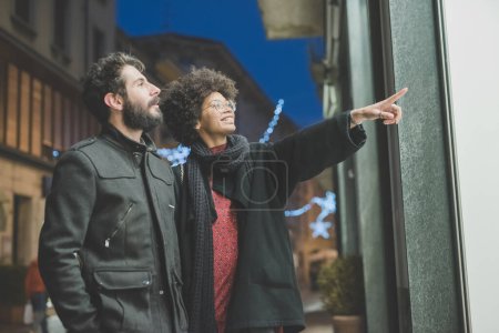 Photo for Young multiethnic couple outdoors at night pointing at shop - Royalty Free Image