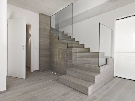 Téléchargez les photos : Gros plan d'un escalier moderne en bois avec une balustrade en verre dans le salon. Le plancher en bois est visible sur la gauche, et il y a une porte blanche ouverte. - en image libre de droit