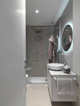 Téléchargez les photos : Salle de bain moderne intérieur au premier plan sur la droite une armoire lavabo avec lavabo de comptoir et un miroir rond suspendu sur le mur au-dessus tandis qu'en arrière-plan il y a une cabine de douche en maçonnerie avec porte en verre - en image libre de droit