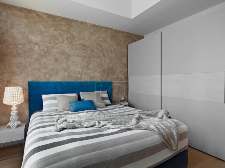 Photo for An interior of the bedroom in the foreground the modern bed in the background on the right a wardrobe with lacquered doors while on the left there is a bedside table with a lit lamp above - Royalty Free Image