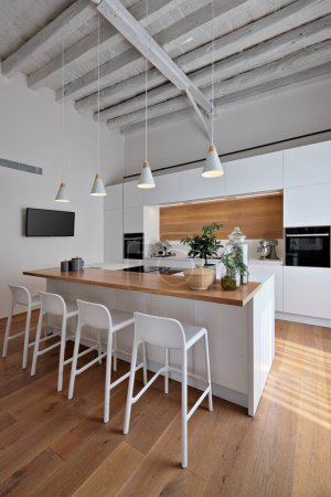 Téléchargez les photos : Vue intérieure d'une cuisine blanche moderne, au premier plan la cuisine îlot avec tabourets, l'éclairage est confié à quatre lampes suspendues, le sol et le plafond sont en bois - en image libre de droit