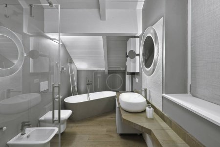 Téléchargez les photos : Vue intérieure d'une salle de bain moderne dans le grenier avec une baignoire autoportante et cabine de douche, à droite le lavabo de comptoir sur le dessus en bois, le sol est en parquet - en image libre de droit