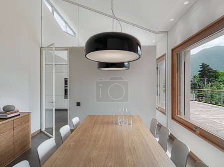 Foto de Primer plano de una gran mesa de comedor moderna con sus sillas y dos lámparas colgantes, en el fondo a la izquierda una puerta de vidrio se abre a la cocina - Imagen libre de derechos