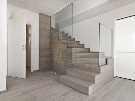 Foto de Primer plano de una moderna escalera de madera con barandilla de vidrio en la sala de estar con suelo de madera a la izquierda hay una puerta blanca abierta - Imagen libre de derechos