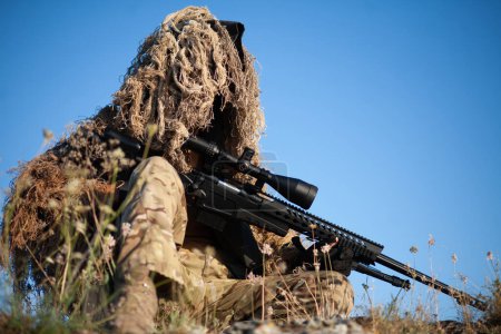 sniper en costume ghillie (camouflage) avec un fusil de haute précision à lunette optique en position de combat en montagne