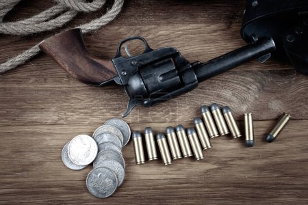 Old west gun with belt, holster and ammunition with silver dollar coins on wooden table