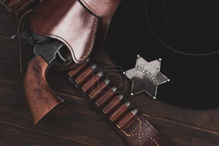 Sheriff star with gun, holster and gun belt with old west black hat on table. Top view.
