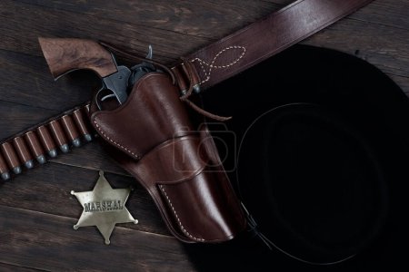 Marshal star badge with gun, holster and gun belt with old west black hat on table. Top view.