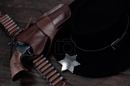 Sheriff star badge with gun, holster and gun belt with old west black hat on table. Top view.