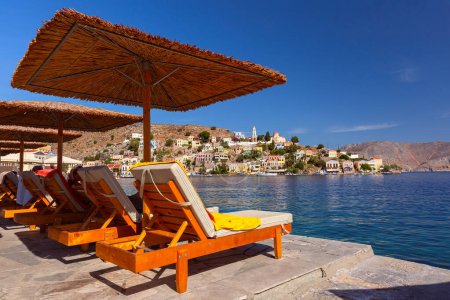 Luxuriöse Liegestühle unter Schilfschirmen mit Blick auf den malerischen Hafen der Insel Symi, Griechenland